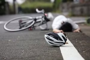 A bicycle and helmet lie on the road as a person is seen lying face down in the background.