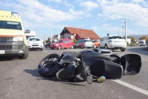 A fallen motorcycle lies in the middle of a road surrounded by several cars and an ambulance under a partly cloudy sky.