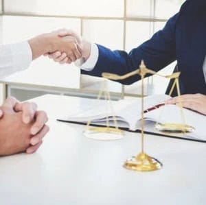 Two people shaking hands across a desk with a justice scale, open book, and clasped hands visible in the foreground.