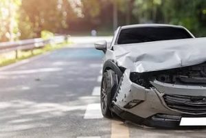 A silver car with significant front-end damage is parked partially on a roadside, surrounded by trees in the background.