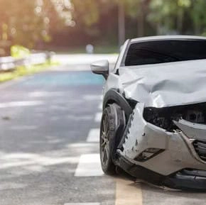 A silver car with significant front-end damage is parked on the side of an empty road with trees in the background.