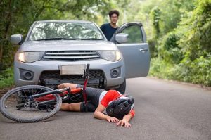 A cyclist lies on the road near a bicycle in front of a car. A person stands by the open car door in a wooded area.