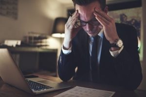 A man in a suit sits at a desk with a laptop, holding his head in his hands and looking stressed while reading a document.