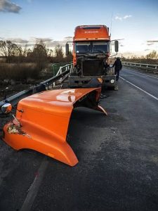 An orange truck with a detached hood on a road, near a bridge. A person stands nearby. Sky is partly cloudy.
