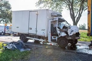 White delivery truck with heavily damaged front after a collision on a rural road, debris scattered around, with emergency vehicle in background.
