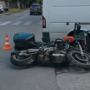 A motorcycle lies on its side next to a white van on a street, with an orange traffic cone nearby.