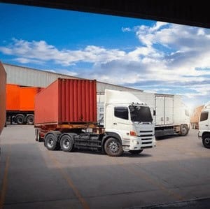 A white semi-truck with a red shipping container is parked near a warehouse, with other trucks and trailers visible in the background under a partly cloudy sky.