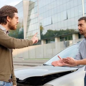 Two men standing beside a car with a damaged hood, engaged in a heated argument in front of a modern glass building.