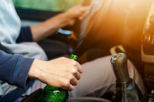 Person holding a green bottle while sitting in the driver’s seat of a car, with hands on the steering wheel and gear shift.