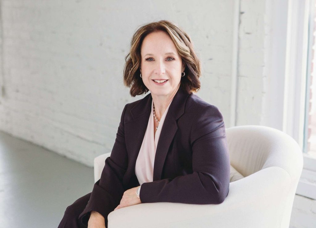 A woman with shoulder-length brown hair in a dark blazer sits in a white armchair, smiling, with a white brick wall and window in the background.