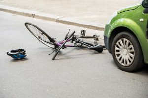 A bicycle lies on the road next to a green car, with a helmet nearby, suggesting an accident.