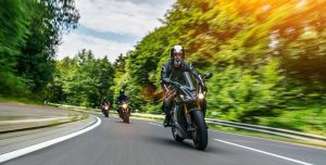 Several motorcyclists ride on a curved road through a lush green forest on a sunny day.