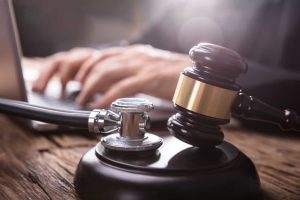 Close-up of a stethoscope and gavel on a wooden surface with a person typing on a laptop in the background.