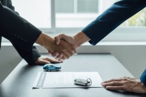 Two people in suits shake hands over a contract on a table, with a car key and a small toy car visible next to the document.