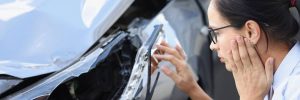 A woman with glasses examines damage to a car's front bumper, holding her face with one hand.