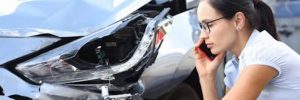 A woman on her phone kneels beside a car with a damaged front bumper and headlight after an accident.