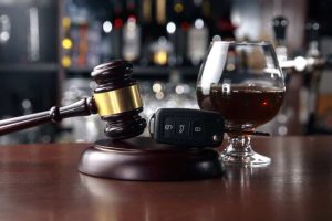 Gavel and car key on a table next to a glass of dark liquor, with blurred shelves of bottles in the background.