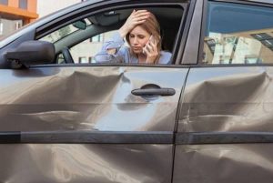 A woman in a parked car, holding her head and speaking on the phone, observes a large dent on the car door.