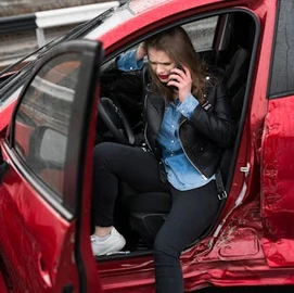 A woman sits in a red car with a damaged door, holding her head and talking on her phone, appearing distressed after an accident.