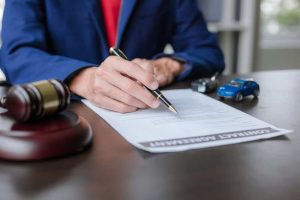 A person in a blue jacket signs a contract at a desk with a gavel and toy cars.
