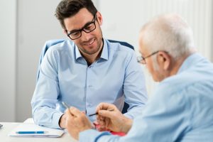 A young man in glasses and a collared shirt smiles while talking to an older man with glasses holding a pen. They are sitting at a table with notebooks and papers.