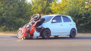 A red motorcycle is overturned on top of a small white car parked on the roadside, with greenery in the background.