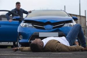 A man lies on the ground in front of a blue car with a damaged hood, while another man stands by the open driver's door.