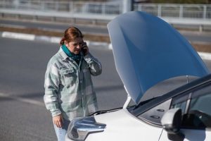 Person standing by a white car with the hood open, talking on a cellphone, on the side of a road.