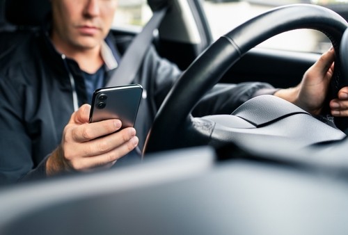 A person sitting in the driver's seat of a car holds a smartphone in one hand while steering with the other.