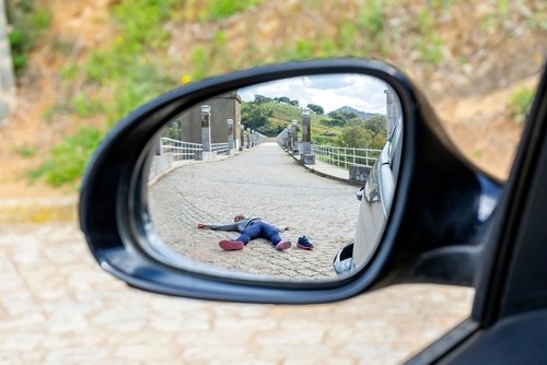 A person is lying on a cobblestone road, seen through the side mirror of a parked car.