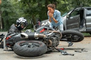 A motorcyclist sits by a fallen motorcycle on the road while a concerned woman stands nearby; a car with an open door is in the background.