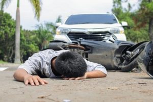 A person lies face down on the road in front of a fallen motorcycle and a white car, suggesting a traffic accident.
