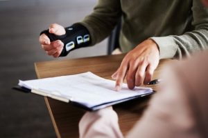 Two people sit at a table; one wears a wrist brace and holds a pen while the other points to a document on a clipboard.