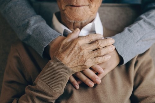 An elderly person sits with another person's arms gently wrapped around their shoulders, both hands resting on the elder's chest.