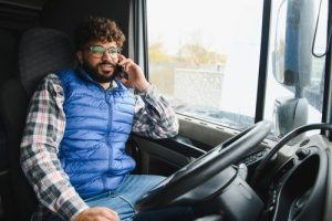 A man sitting in a truck cab, wearing glasses, a blue puffer vest, and plaid shirt, talking on a smartphone.
