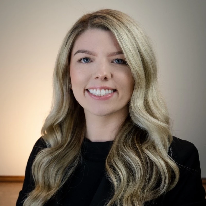 A woman with long, wavy blonde hair and a black top smiles at the camera in front of a plain light background.