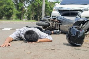 A person lies face down on the road near a fallen motorcycle and a car; a helmet is on the ground nearby.