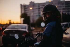 A person wearing a helmet sits on a motorcycle in city traffic at dusk, with cars and a building visible in the background.