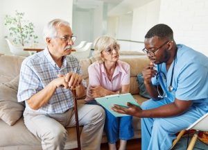 A healthcare professional in scrubs shows a clipboard to an older man and woman sitting on a couch, discussing medical information.