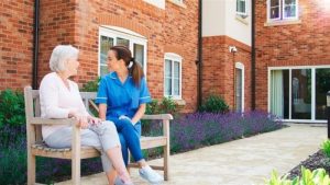An elderly woman and a caregiver in scrubs sit on a wooden bench outside a brick residential building, talking and smiling.