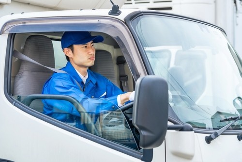 A man in a blue uniform and cap is sitting in the driver’s seat of a white truck, looking forward with his hands on the steering wheel.