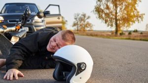 A person lies on the road in pain next to a fallen motorcycle and helmet, with a car parked nearby and trees in the background.