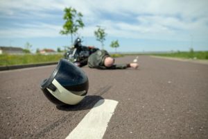 A motorcycle helmet lies on the road with a person and a motorcycle in the background, suggesting a recent accident.