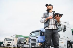 A man standing in front of parked semi-trucks holds a tablet, wearing a cap, vest, and jeans, looking to the side outdoors.
