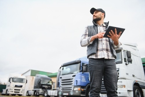bearded truck driver in vest and cap holding tablet near commercial trucks