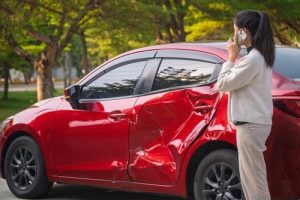 A woman stands by a red car with a large dent in the side, talking on her phone, possibly reporting the accident.