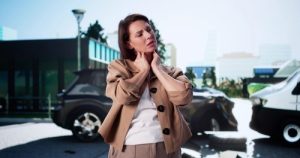 A woman stands outdoors holding her neck, with two damaged vehicles in the background, suggesting a recent car accident.