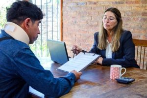 A man wearing a neck brace reviews documents with a woman at a desk; a laptop, mug, and calculator are also on the table.