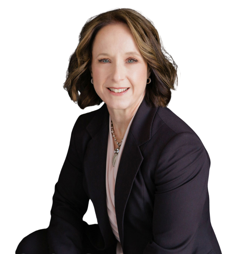 A woman with shoulder-length brown hair wearing a dark blazer, light blouse, and necklace sits and smiles at the camera against a plain white background.