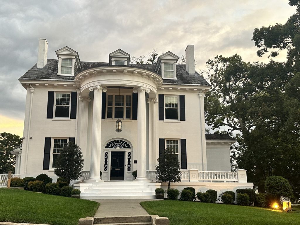 Large white colonial-style house with black shutters, four tall columns at the entrance, manicured bushes, and a wide front lawn under a cloudy sky.
